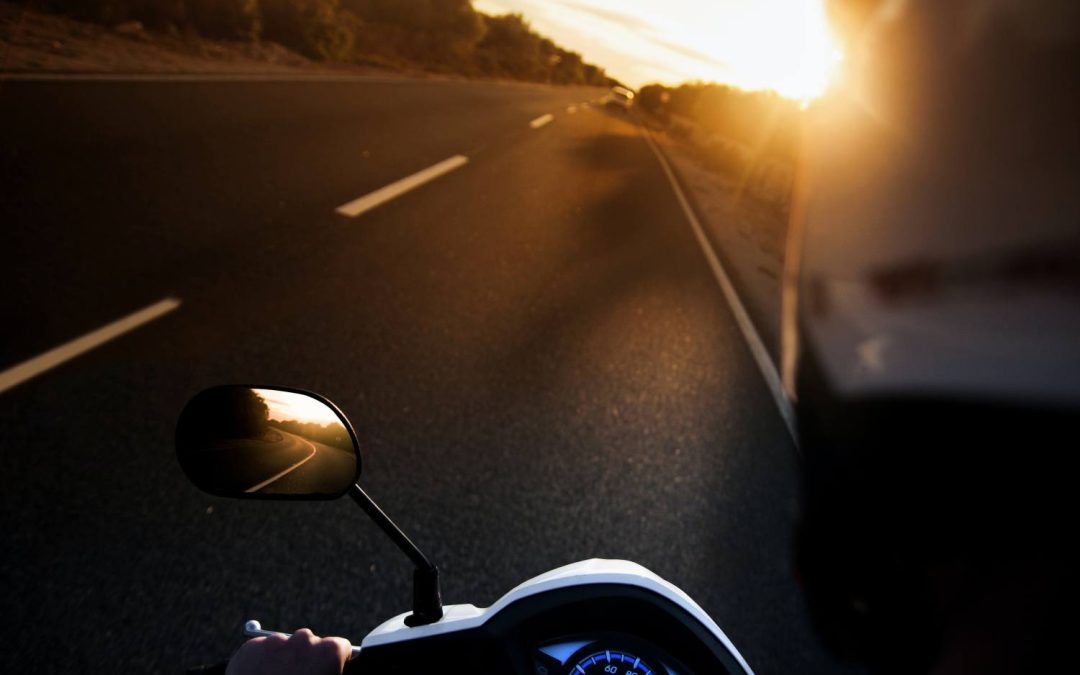 Motorcycle Training Blog Motorbike rider's perspective on an open road during sunset, showcasing the motorcycle's dashboard and side mirror reflecting the winding path ahead.