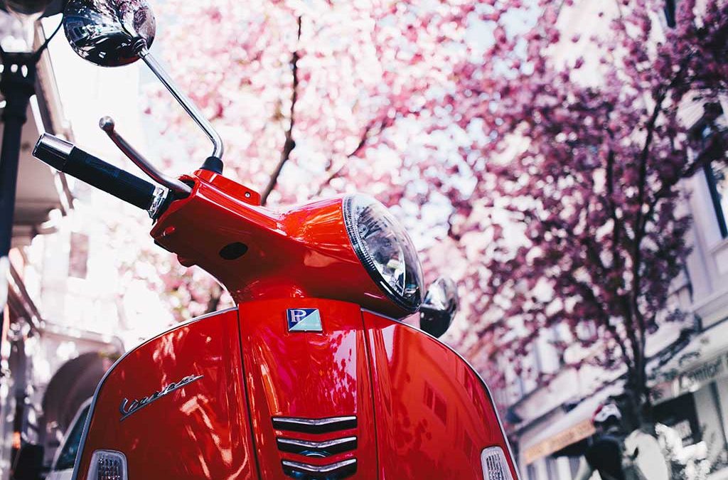 Motorcycle Training Blog Red Vespa scooter parked under blooming cherry blossom trees, symbolizing motorcycle training and riding culture.