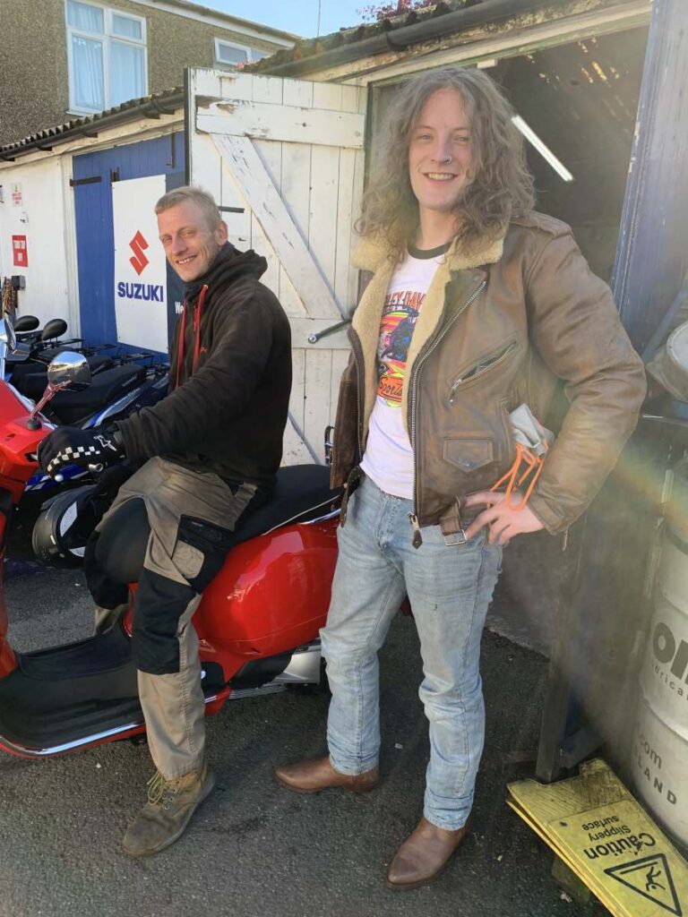 Two motorcycle instructors smiling, one seated on a red Lexmoto scooter, the other standing beside him in a leather jacket, showcasing the friendly atmosphere of North London Motorcycle Training.