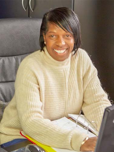 Sandra Yally, office team member at North London Motorcycle Training, smiling in a cozy sweater at her desk, emphasizing dedication to motorcycle training.