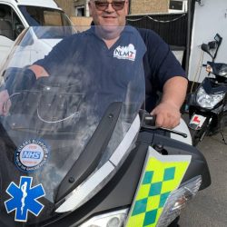 Man smiling while seated on a Lexmoto motorcycle, wearing a North London Motorcycle Training (NLMT) polo shirt, with visible NHS and emergency service logos, showcasing the motorcycle training environment.