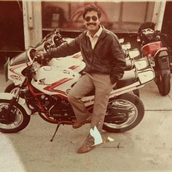Person sitting on a Lexmoto motorcycle, wearing a leather jacket, with a confident expression, surrounded by additional motorcycles in a training environment.