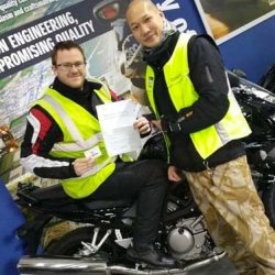 Two motorcycle trainers in high-visibility vests celebrating a student's achievement, holding a certificate, in front of a Lexmoto motorcycle, showcasing North London Motorcycle Training's commitment to rider success.