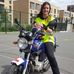 Smiling female motorcycle instructor in high-visibility jacket sitting on a Lexmoto motorcycle, showcasing motorcycle training at North London Motorcycle Training.