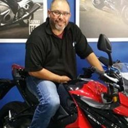 Man sitting on a Lexmoto motorcycle, smiling, in front of motorcycle-themed wall displays, representing the dedicated team at North London Motorcycle Training.