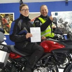 Person seated on Lexmoto motorcycle holding training certificate, smiling instructor giving a thumbs-up, motorcycle training environment.